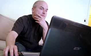 Ove Villadsen sidder i en sort t-shirt i sin sofa foran en bærbare computer. 