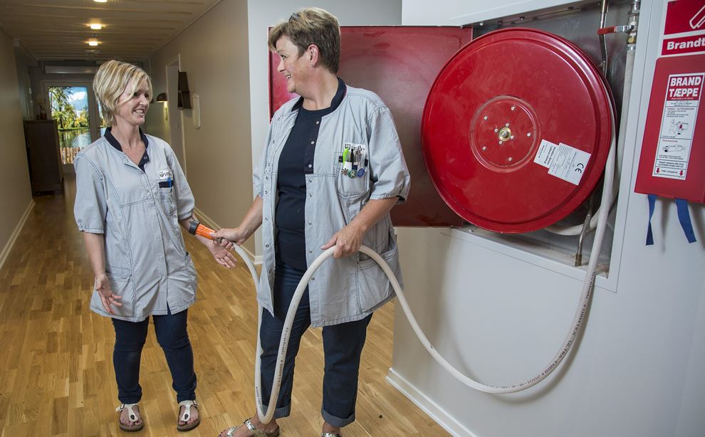 To kvindlige sosu-assistenter står på en gang på et plejehjem og tale sammen. Den ene holder en brandslange i hånden.