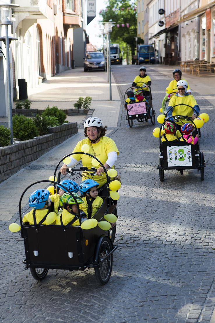 Fire voksne kører på fire cykler, alle iført gult tøj. foran i en boks på cyklen sidder en masse børn, også iført gult tøj