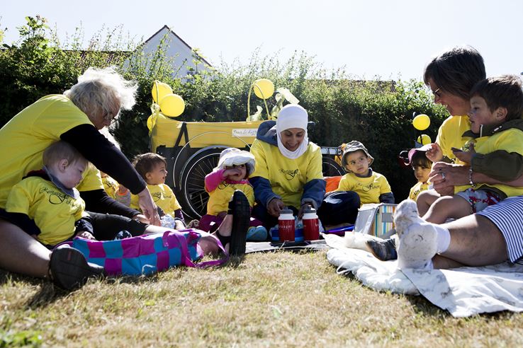 En masse voksne og børn sidder på nogle tæpper på en græsplæne, alle iført gule bluser