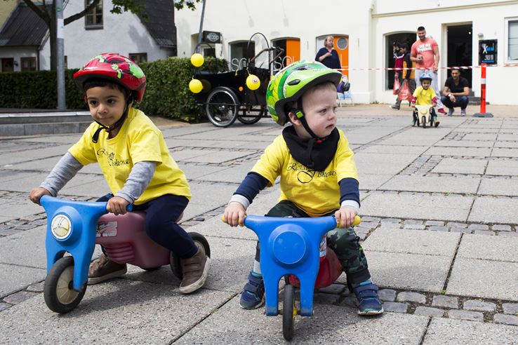 To børn sidder på to små blå cykler på et fortov, begge iført gule bluser og farvede cykelhjelme