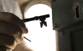 En hånd er ved at sætte en gammel nøgle ind i en gammel lås - bagved ser man et stort buet vindue uden ruder