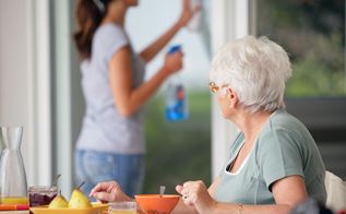Senior kvinde har morgenmad med en kvinde fra hjemmeplejen i baggrunden, som er i gang med at vaske en rude