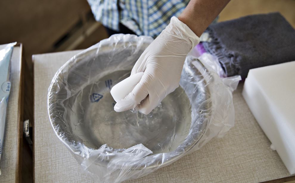En hånd iført en hvid plastic handske dypper en klud i en vand skål der er pakket ind i plastic