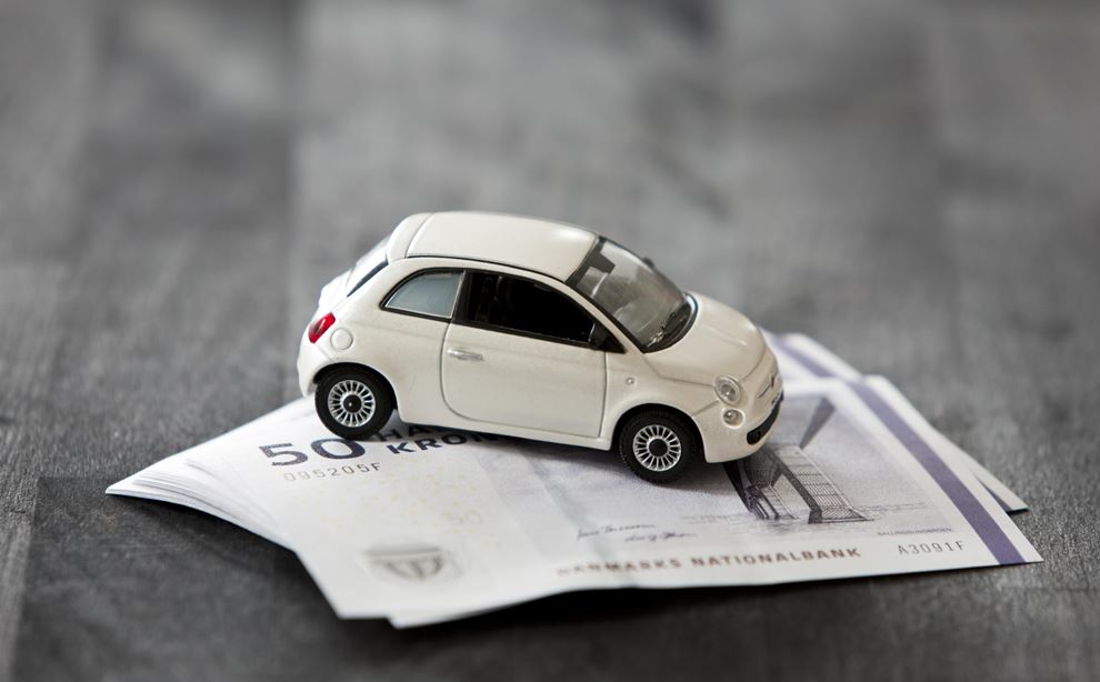 En hvid legetøjs bil står ovenpå et bundt 50-krone sedler, som igen er placeret på et trægulv. 