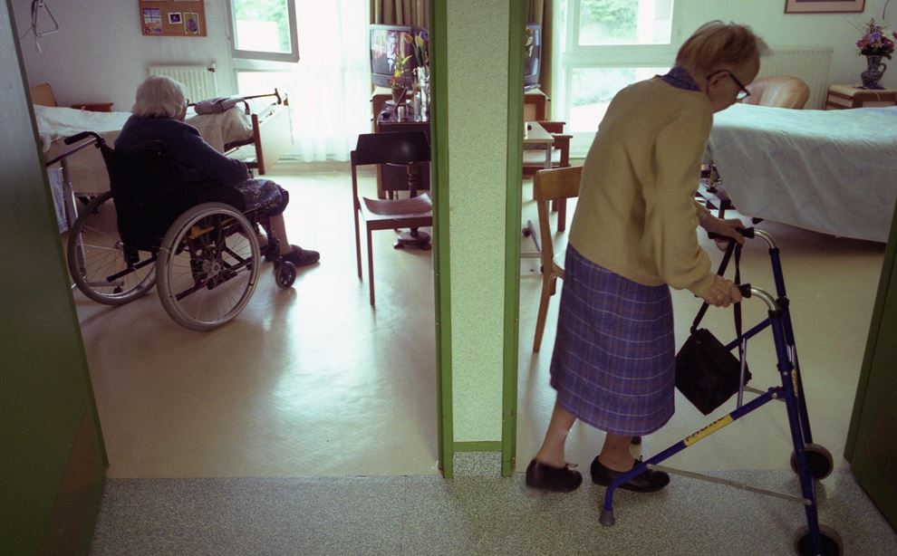 En ældre kvinde i kørestol sidder på sit værelse, mens en anden ældre kvinde går forbi med rollator