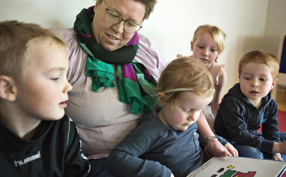 Anna sidder, iført stribet bluse og farvet halstørklæde, sammen med fire børn og læser i en bog