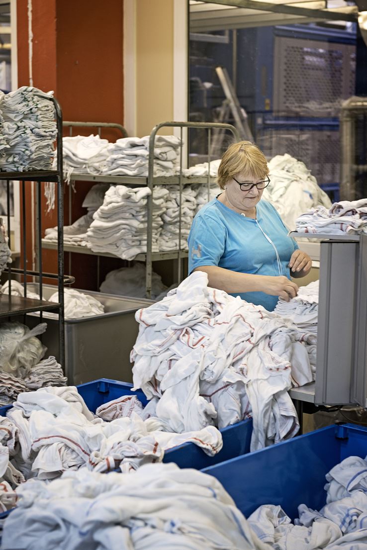 En kvinde står og lægger en masse tøj sammen, omkring hende ligger der hvide uniformer i mange bunker