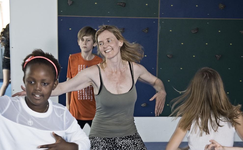 En kvinde står og danser, mens der står tre børn og ligeledes danser omkring hende. Bagved hende står en dreng i orange trøje stille. 