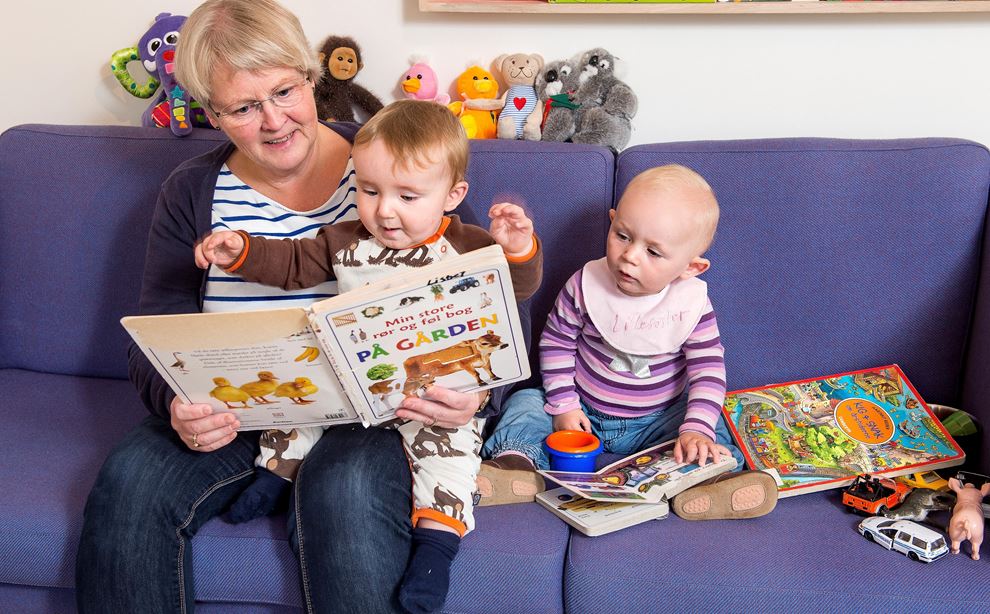 Dagplejer sidder i sofa med to dagplejebørn og læser historie 