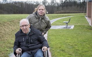En ældre mand med gråt hår sidder i en kørestol, mens en yngre kvinde med lyst hår skubber kørestolen.