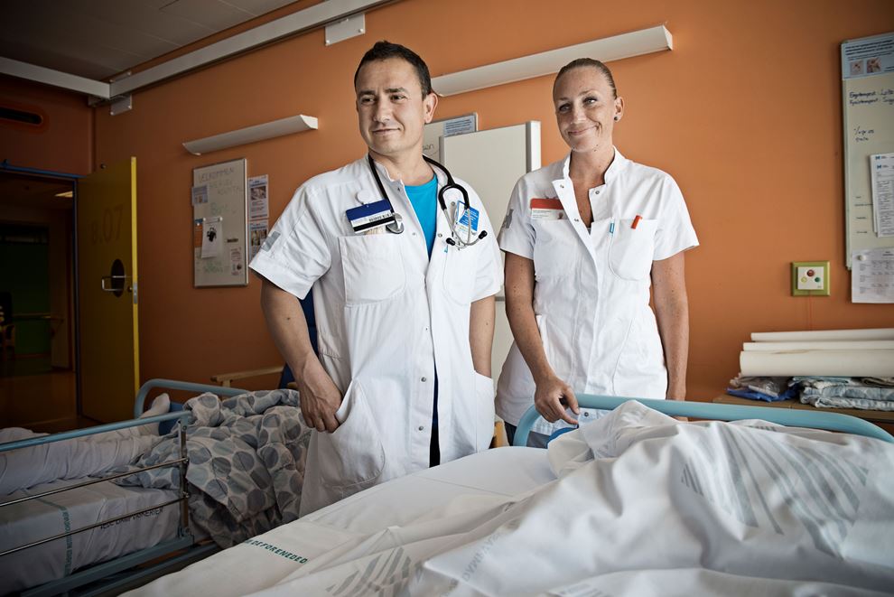 En mand og en kvinde i hvide kitler står ved en sygeseng og kigger op mod hovedgærdet.