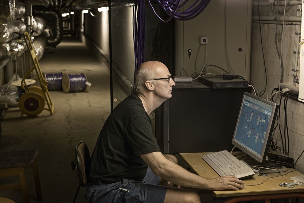 En mand, iført sort t-shirt og briller, sidder i en lang kælder og kigger på en computerskærm