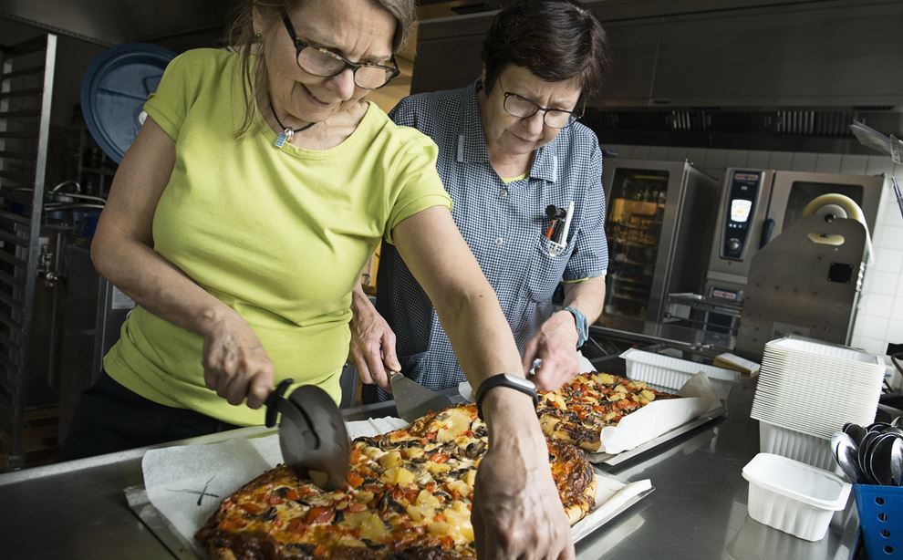 En kvinde i grøn bluse står og skær i en pizza. Ved siden af hende står en kollega i blå trøje og hjælper hende.