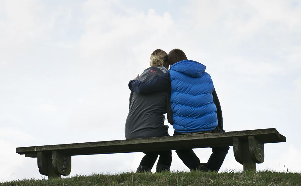 En mand og en dame sidder på en bænk, med ryggen til kameraet og kigger op på himlen. Manden er iført blå dunjakke og damen bærer en grå hættetrøje