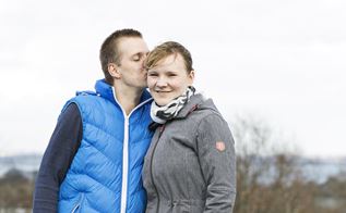 En mand kysser en kvinder på siden af hovedet, mens de står på et bakketop med en park og fodboldbane i baggrunden
