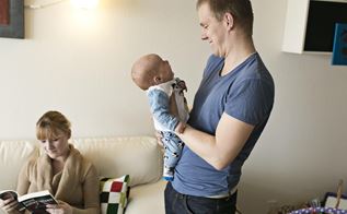 En mand står med et spædbarn i sine arme, mens der sidder en kvinde på sofaen bag ham og læser i en bog