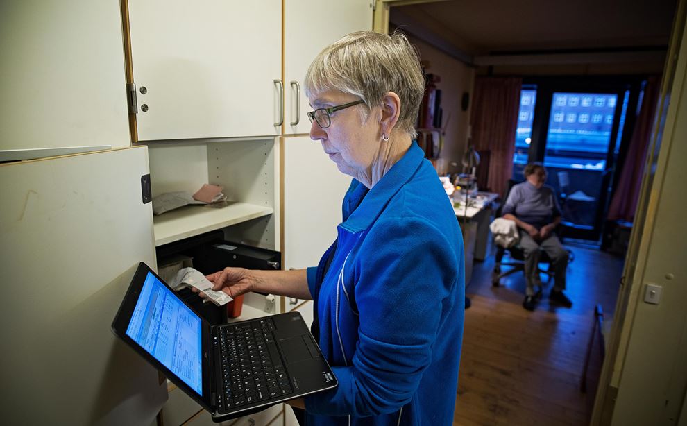 Kvinde i blå trøje og med briller står og kigger på en computerskærm ved indgangen til en ældrebolig.