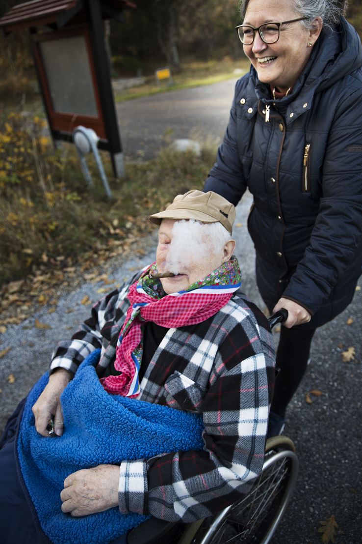 En ældre mand bliver kørt i kørestol af en kvinde, imens han ryger på en cigar