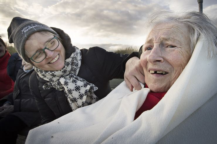 En ældre kvinde sidder med et tæppe op over ørene, mens en yngre kvinde sidder ved siden af og kigger smilende over på hende