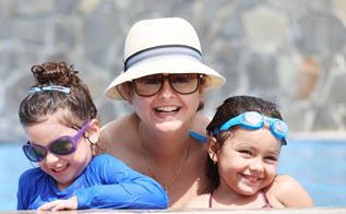 En kvinde iført solhat og solbriller sidder i en swimmingpool med to børn.
