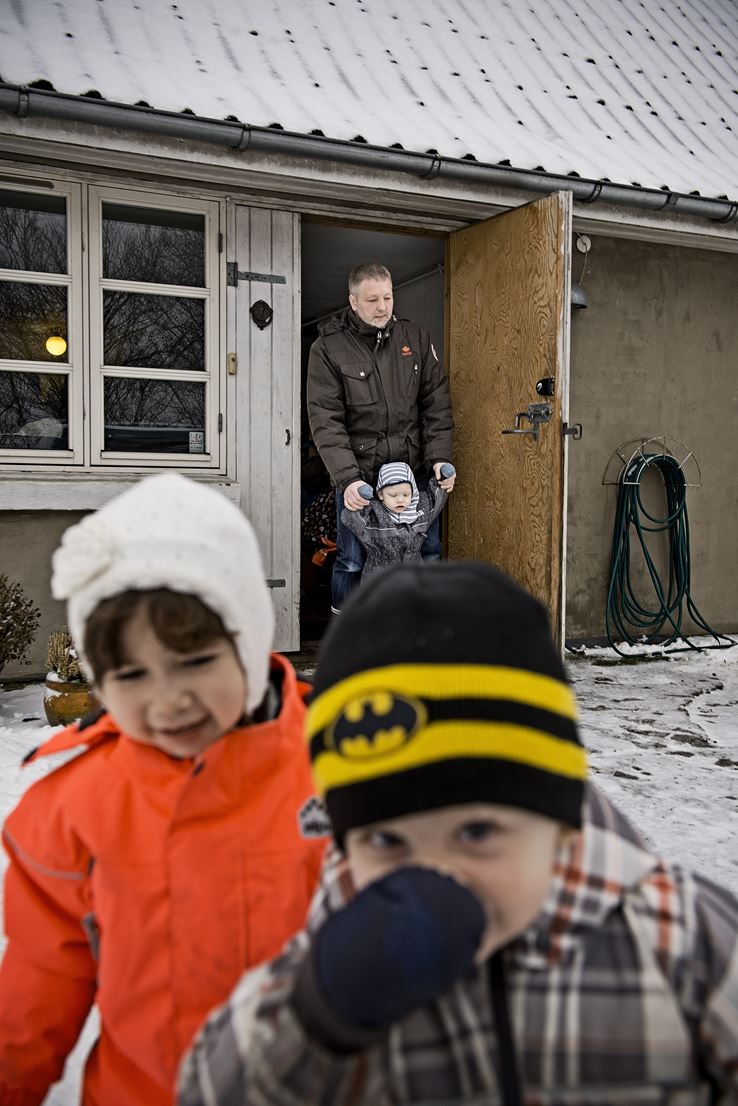 To børn i forgrunden, iklædt vintertøj med hue og vanter. I baggrunden kommer Kim ud af et hus med et andet barn, som han holder i hænderne.