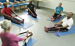 Fem mennesker sidder på gulvet ovenpå nogle yoga-måtter, hvor de strækker ud med et plastic bånd