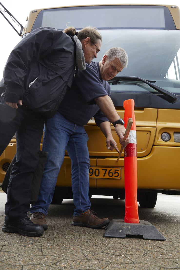To mænd står med et målebånd og måler afstanden mellem en rød trafikkegle og en gul bus