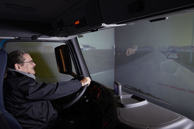 En mand sidder i en lastbilssimulator, hvor ruderne er skiftet ud med skærme som simulerer at han kører ude på en vej