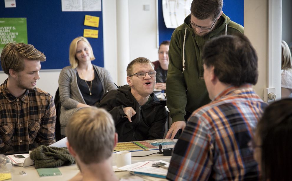 Elever sidder i et klasseværelse. En spastisk lammet mand sidder i midten, mens en ung mand i grøn trøje står op.