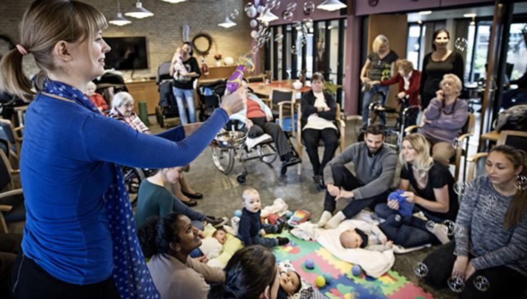 En kvinde iført en blå bluse blæser sæbebobler ud over en gruppe forældre og babyer, der ligger på gulvet.