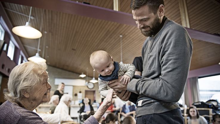 En mand med grå trøje holder en baby, mens en kvinde med gråt hår kigger på-