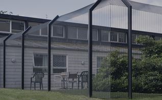 En bygning med et sort hegn rundt om. der står nogle havemøbler indenfor hegnet.
