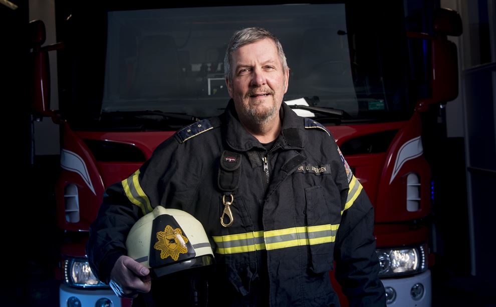 En brandmand står i uniform foran en brandbil med sin hjelm i højre hånd