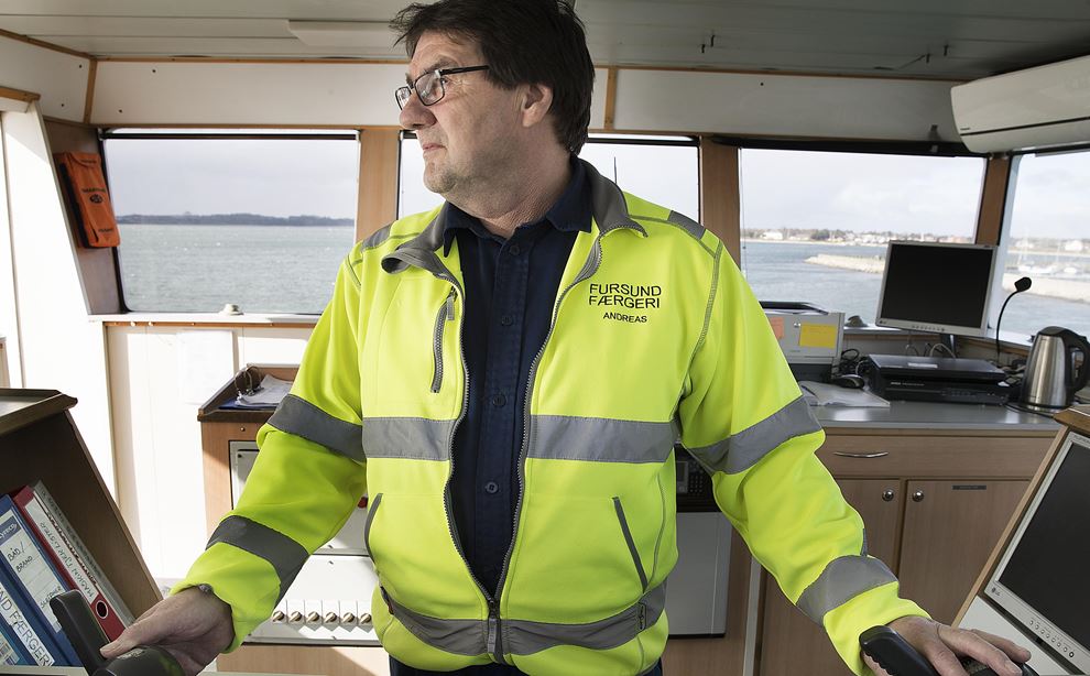 En mand i en gul jakke med reflekser på står i et styrhus på en færge. Han står og styrer med to joysticks.