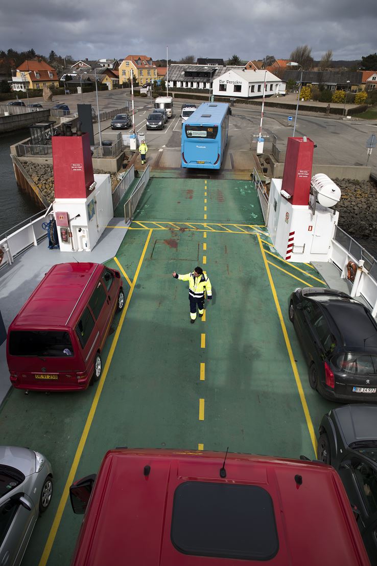 Et grønt færgedæk, hvor der står en mand i gul jakke og peger, hvor bilerne skal køre hen. Der er en blå bus der er ved at bakke ombord på færgen.