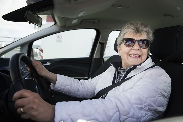 En kvinder sidder i en bil med en hvid jakke på og holder begge hænder på rattet. Kvinden smiler og har solbriller på.