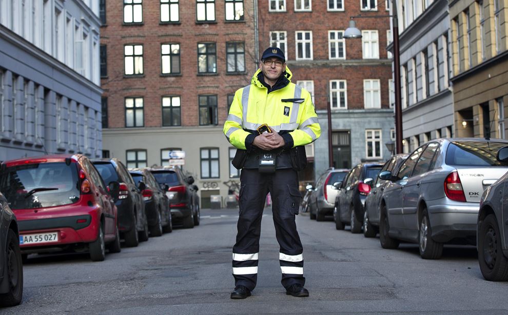 En mand - iført uniform - står midt på en vej. På hver side af vejen er der parkeret biler