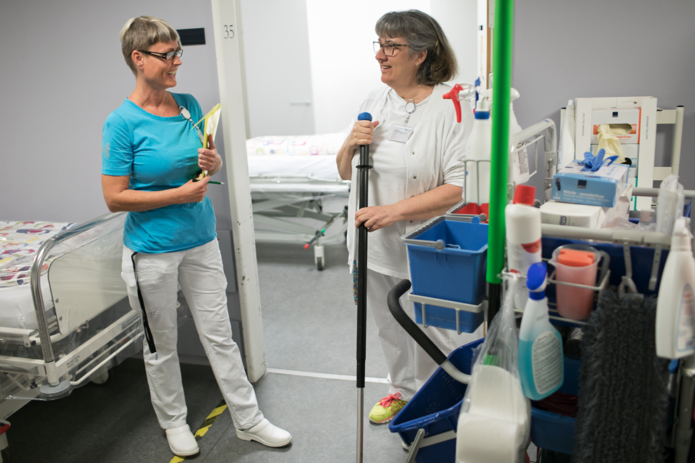 To kvinder står på et hospitalsværelse, den ene er iført rengøringsuniform og står med en støvsuger i hånden