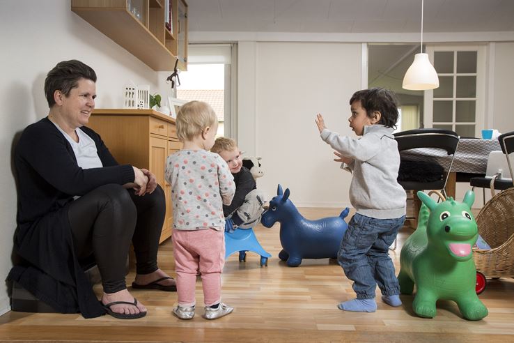 En kvinde sidder på gulvet og smiler, mens der står tre børn ved siden af hende og underholder