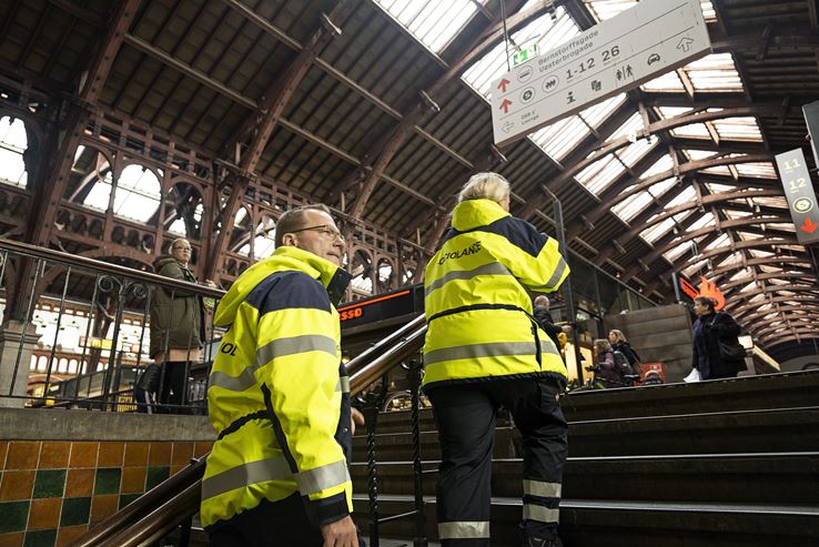 To mennesker, iført gule uniformer, går op ad en trappe til en stor stationshal