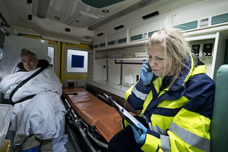 En kvinde sidder, iført gul uniform, i en ambulance, mens der sidder en mand iført tæppe ved siden af hende