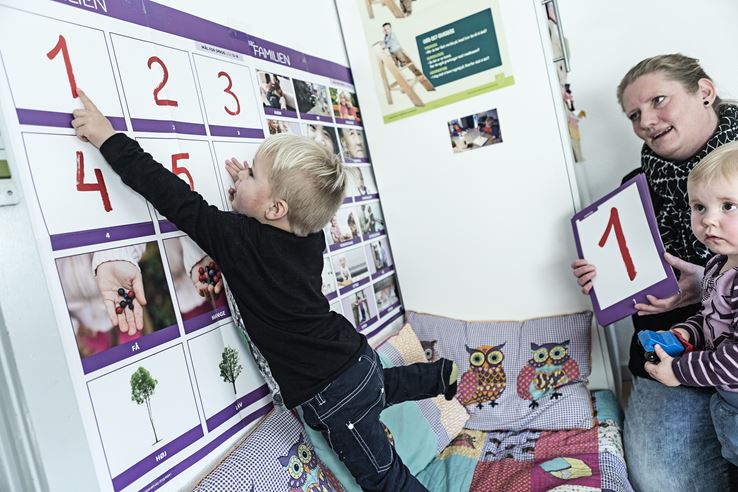 En barn peger op på et rødt et-tal som hænger på væggen, mens en kvinde og et andet barn sidder med et andet et-tal og kigger på