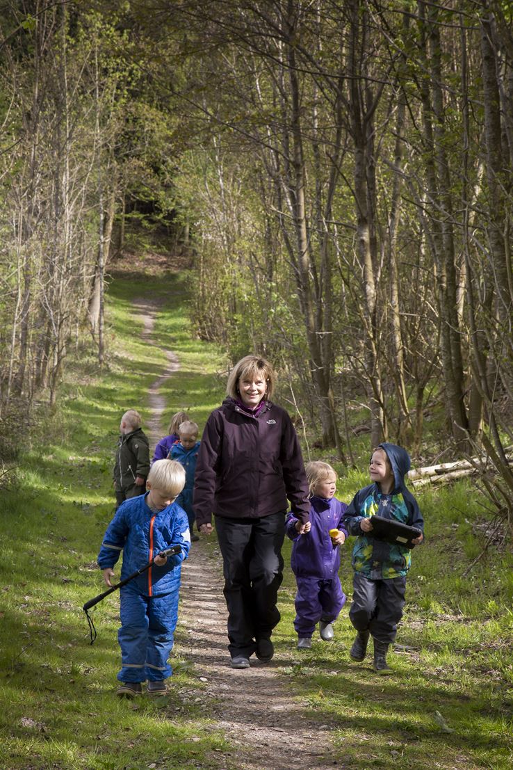 En kvinde går på en sti i en skov. Omkring hende går seks børn. 