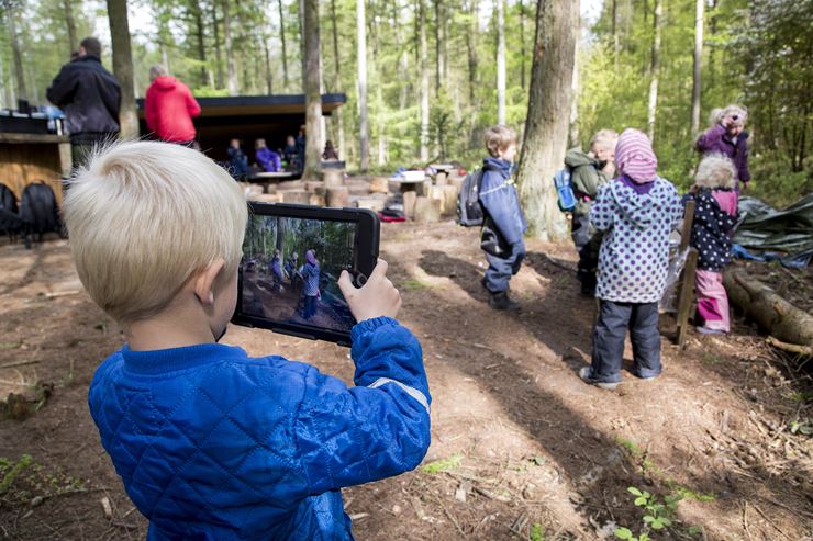 En dreng står med en iPad i en skov, mens der står en masse andre børn og vokse omkring ham