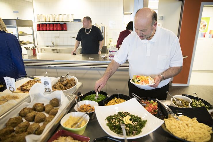 En mand, iført sorte bukser og hvid skjorte, står og tager mad fra en buffet i en kantine