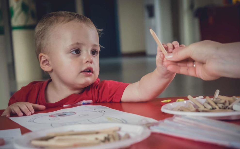 Barn sidder ved et bord og tegner, mens en voksen rækker barnet en blyant.