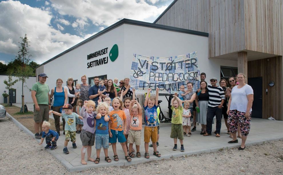 En stor gruppe af børn og voksne står foran en hvid bygning, mens nogle af de voksne bærer et banner med skrift.