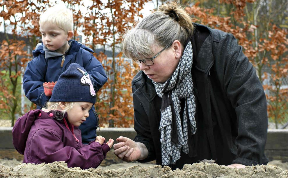 Kvinde med sort jakke og opsat hår sidder ved i sandkasse sammen med to børn