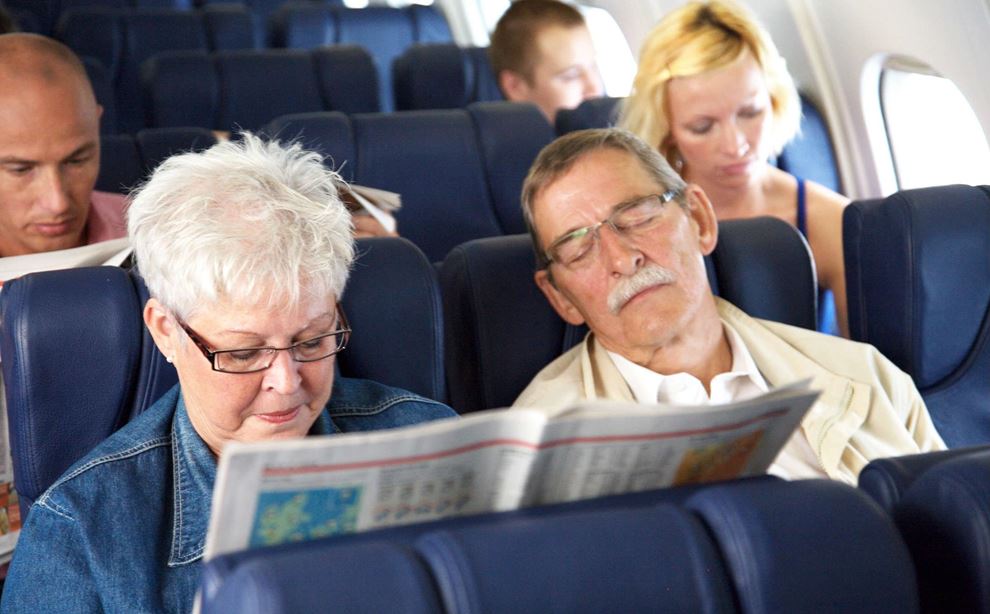Et par sidder på flyet på vej på ferie. Kvinden læser avis, mens manden sover.